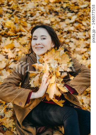 Smiling woman throws autumn leaves in park while looking at camera. Finding joy in nature, stress reduction techniques, emotional health awareness. Smiling woman throws autumn leaves in park while looking at camera. Finding joy in nature, stress reduction techniques, emotional health awareness. 140077558