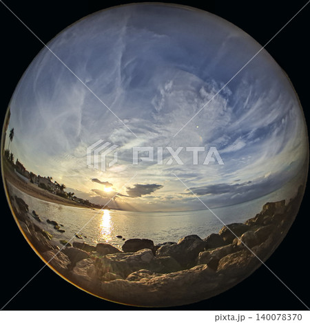 Sunset occurs over the sea with sun rays breaking through clouds. Waves gently lap against the rocks along the shore. The sky is filled with colors and formations from the clouds. 140078370