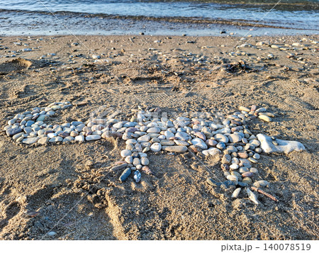 On a sandy beach near the water, a large mosaic of a lizard is made entirely of smooth sea pebbles. The art piece displays careful arrangement of stones under clear skies. On a sandy beach near the water, a large mosaic of a lizard is made entirely of smooth sea pebbles. The art piece displays careful arrangement of stones under clear skies. 140078519