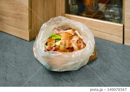 Trash bag filled with vegetable scraps sits by the kitchen cabinet in the afternoon light 140078567
