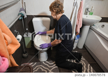 Woman cleaning toilet in a small bathroom during daytime hours Woman cleaning toilet in a small bathroom during daytime hours 140078578