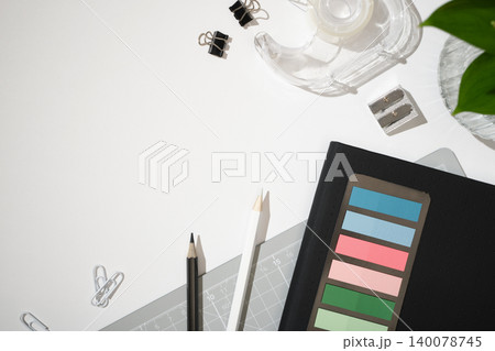 Top view of office supplies on a white desk. Black notebook with colorful sticky tabs, pencils, a cutting mat, tape dispenser, and paper clips. Copyspace Top view of office supplies on a white desk. Black notebook with colorful sticky tabs, pencils, a cutting mat, tape dispenser, and paper clips. Copyspace 140078745