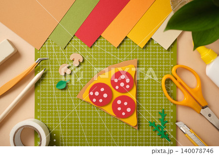 Flat lay of a paper pizza slice with pepperoni on a green cutting mat. Creative DIY workspace with colorful paper sheets, scissors, glue, tweezers, and stationery for art and craft projects. Flat lay of a paper pizza slice with pepperoni on a green cutting mat. Creative DIY workspace with colorful paper sheets, scissors, glue, tweezers, and stationery for art and craft projects. 140078746