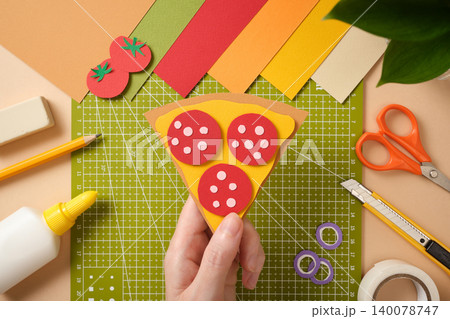 Hand is holding a finished paper craft: a slice of pepperoni pizza. There are paper craft supplies around it. Hand is holding a finished paper craft: a slice of pepperoni pizza. There are paper craft supplies around it. 140078747