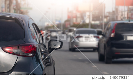 Close-up rear view of a dark grey hatchback waiting in a dense daytime traffic jam. A blurry line of various commuter cars can be seen stretching down the urban street ahead. Close-up rear view of a dark grey hatchback waiting in a dense daytime traffic jam. A blurry line of various commuter cars can be seen stretching down the urban street ahead. 140079334