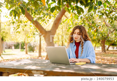 Attractive woman working or studying at laptop as sitting in city park. Freelancer working in park. 140079953