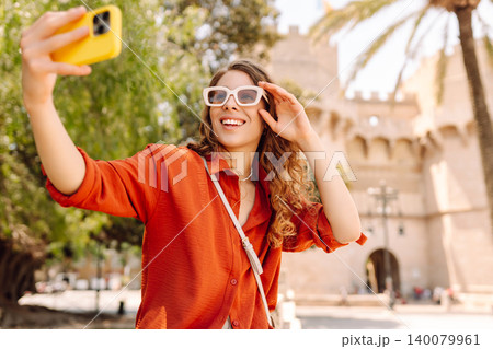 Cheerful woman taking a selfie in stylish sunglasses while strolling down a sunlit city street 140079961