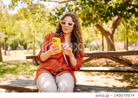 Young woman sits on bench in park, using smartphone while dressed casually. Travel, blogging concept 140079974