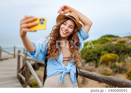 Young woman with curly hair wears a stylish hat and waves while taking a selfie. Travel, tourism 140080091