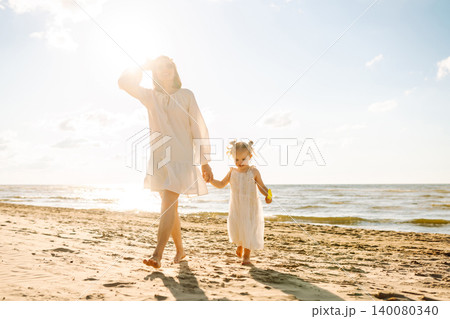 A mother and daughter enjoy a sunny beach stroll, as waves gently lap at their feet during sunset A mother and daughter enjoy a sunny beach stroll, as waves gently lap at their feet during sunset 140080340