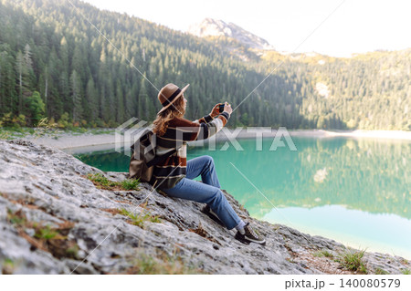 Young woman takes a photos the nice view by the serene lake surrounded by forested mountains 140080579