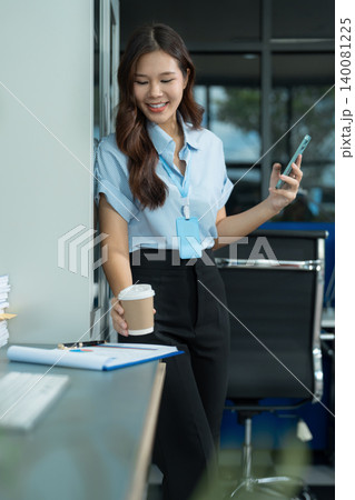 Asian businesswoman sits in a office working on smartphone and enjoys a coffee. Asian businesswoman sits in a office working on smartphone and enjoys a coffee. 140081225