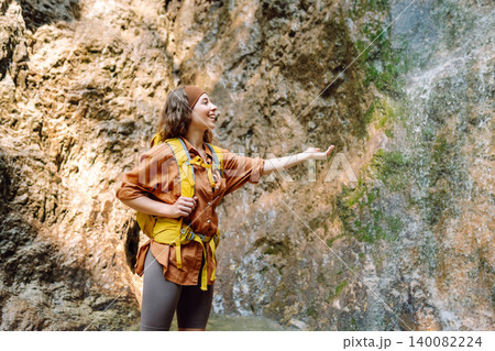 Smiling female dressed in hiking clothes, enjoys a waterfall near a mountain river. Travel, trekking 140082224
