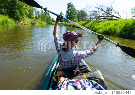 Woman raises paddle above head while kayaking, river trip, active water sport, summer adventure, energetic outdoor lifestyle, tourist use. 140085443