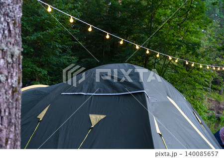 Tent with garland in wooded campsite, outdoor travel area, nature tourism, festive camping application and group adventure recreation. Tent with garland in wooded campsite, outdoor travel area, nature tourism, festive camping application and group adventure recreation. 140085657