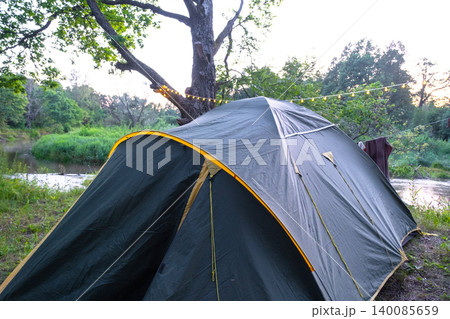 Classic dome tent in green grassy area under tree, tranquil outdoor camping, tourism, rural landscape, adventure travel application. 140085659