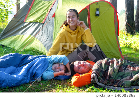 Mother and two daughters in sleeping bags, tent nearby, family picnic in nature, sportswear and outdoor gear. Mother and two daughters in sleeping bags, tent nearby, family picnic in nature, sportswear and outdoor gear. 140085868