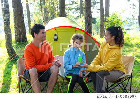 Family sitting together in camping chairs, mother, father and daughter, using mosquito spray, safe child, protection from allergy, healthy outdoor fun, summer vacation. 140085907