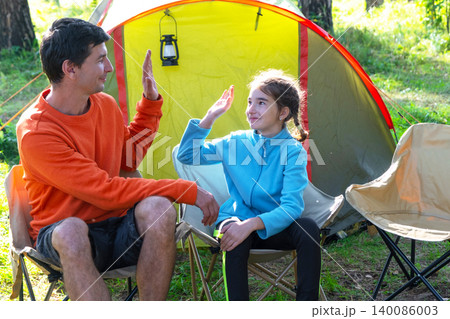 Cheerful family moment by camping tent: dad, daughter and mom, comfy nature site, vacation and sports attire. Cheerful family moment by camping tent: dad, daughter and mom, comfy nature site, vacation and sports attire. 140086003