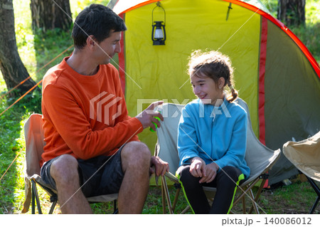 Father holding mosquito repellent while daughter observes, family at campsite with camping gear, emphasizing safety, allergy risks, disease prevention, summer care. 140086012