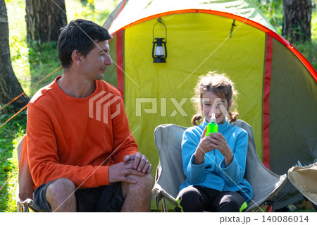 Smiling daughter sitting in camping chair, father nearby, family camping with mosquito spray, safe child, allergy and disease prevention, joyful outdoor lifestyle. Smiling daughter sitting in camping chair, father nearby, family camping with mosquito spray, safe child, allergy and disease prevention, joyful outdoor lifestyle. 140086014