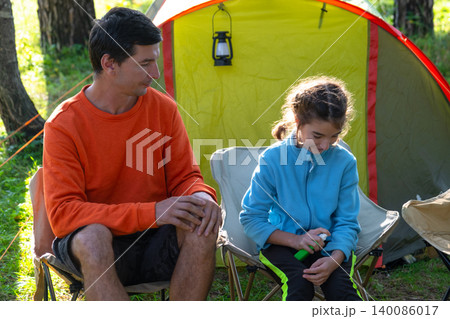 Father handing mosquito repellent to daughter, child protected outdoors, family by tent, allergy and illness prevention, caring emotions, summer adventure. 140086017
