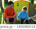 Daughter holding spray bottle, father watching, family outdoors at campsite, mosquito protection, bite allergy, summer health care, parent and child bonding. 140086018
