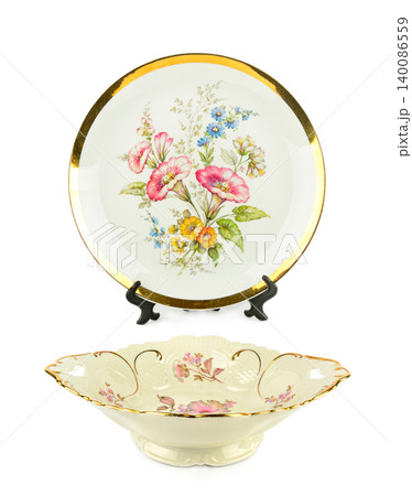 Elegant vintage porcelain plate with gold rim on a stand next to a decorative ceramic bowl with painted floral patterns isolated on a white background. Elegant vintage porcelain plate with gold rim on a stand next to a decorative ceramic bowl with painted floral patterns isolated on a white background. 140086559