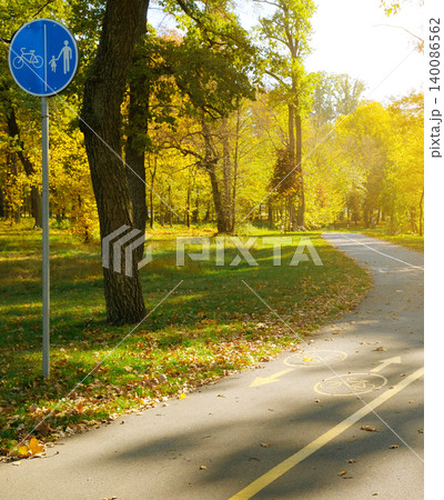 Blue round bicycle and pedestrian shared path sign on a metal pole next to an asphalt track winding through a beautiful public park filled with golden autumn trees. Blue round bicycle and pedestrian shared path sign on a metal pole next to an asphalt track winding through a beautiful public park filled with golden autumn trees. 140086562
