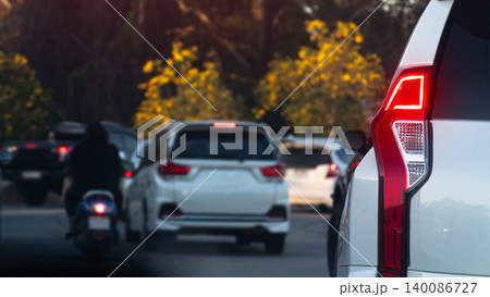 White SUV with its distinctive red tail light is stuck in traffic. Motorcycle rider and a white sedan are visible further ahead on the busy tropical tree with yellow flowers street.  140086727