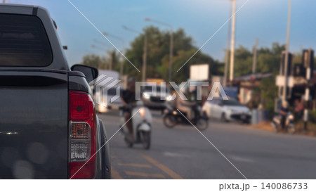 View looking past the rear corner of a dark truck stopped on asphalt road. Several motorcycles and cars navigate through the traffic lanes under the clear afternoon sky. Rural urban background. 140086733