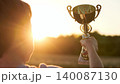 Small victor grips polished chalice against glowing horizon in vivid child victory. Bright silhouette lifts shining cup echoing kid triumph. Soft sunset beams infuse inspiring atmosphere child triumph 140087130