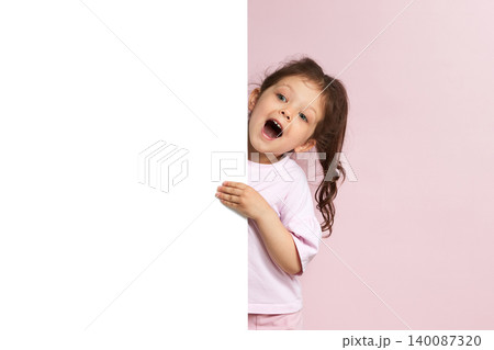 Little girl with wide open mouth peeking from behind a blank white banner with copy space, standing against a pink background Little girl with wide open mouth peeking from behind a blank white banner with copy space, standing against a pink background 140087320