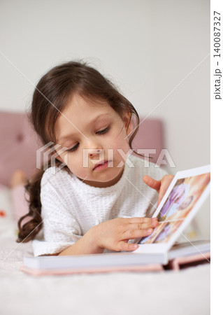 Little girl on bed flipping through family photo album, quietly reminiscing childhood memories and treasured moments at home Little girl on bed flipping through family photo album, quietly reminiscing childhood memories and treasured moments at home 140087327