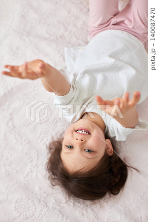 Happy little girl lying upside down on a soft bed, smiling and reaching arms up, enjoying playful childhood moments Happy little girl lying upside down on a soft bed, smiling and reaching arms up, enjoying playful childhood moments 140087470