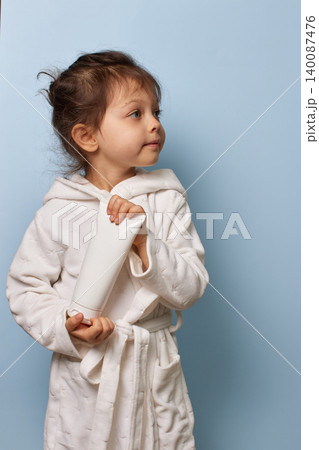 Child girl wearing a white bathrobe holding a blank cream tube on a blue background Child girl wearing a white bathrobe holding a blank cream tube on a blue background 140087476