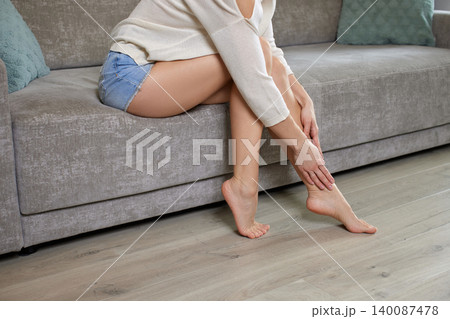 woman sitting on sofa, massaging her tired legs and feet, suffering from pain, swelling, and varicose veins woman sitting on sofa, massaging her tired legs and feet, suffering from pain, swelling, and varicose veins 140087478