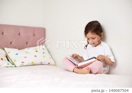 Small child learning and developing literacy skills by reading storybook while sitting on bed at home Small child learning and developing literacy skills by reading storybook while sitting on bed at home 140087504