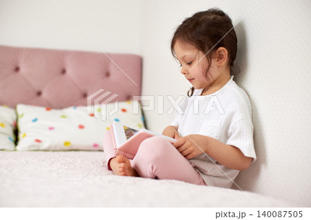 Little child engrossed in a story, turning pages of a book while sitting comfortably on a pink bed at home 140087505