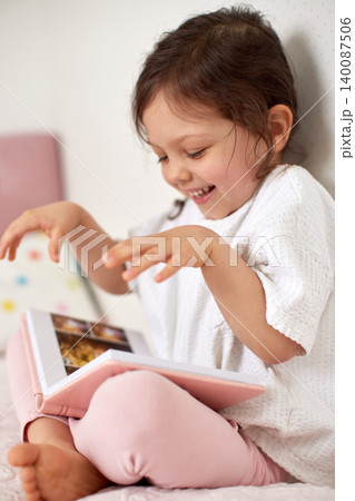 Little child girl sitting on a bed, looking at a photo album and laughing, remembering pleasant childhood moments 140087506