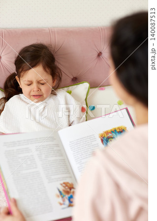 Little girl crying in bed while her mother reads a bedtime story, showing childhood emotions and family moments Little girl crying in bed while her mother reads a bedtime story, showing childhood emotions and family moments 140087513