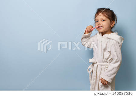 Child girl in white bathrobe practicing brushing teeth against a clean blue background, copy space Child girl in white bathrobe practicing brushing teeth against a clean blue background, copy space 140087530