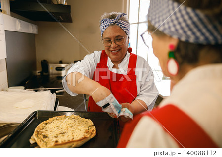 Smiling Mature Hispanic Woman Baking Pizza in a Bright Community Kitchen with a Female Friend Smiling Mature Hispanic Woman Baking Pizza in a Bright Community Kitchen with a Female Friend 140088112