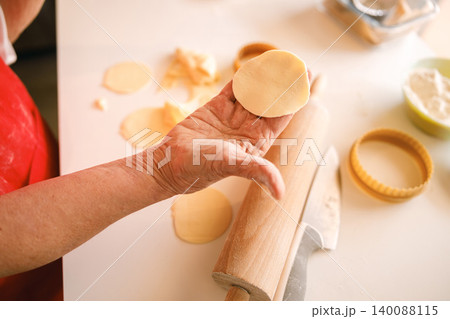 Elderly Caucasian woman hands making circular dough for cookies in a sunlit domestic kitchen Elderly Caucasian woman hands making circular dough for cookies in a sunlit domestic kitchen 140088115
