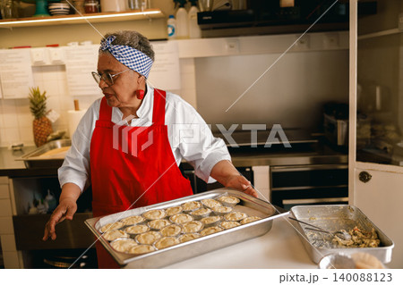Senior Black female chef in red apron holding a tray of mini pies in a professional kitchen environment 140088123
