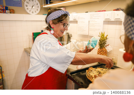 Happy senior Caucasian female chef preparing fresh food on a baking sheet in a sunny indoor kitchen 140088136