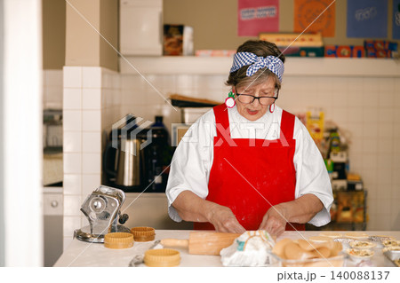 Senior Caucasian woman in red apron preparing artisanal dough in a bright professional kitchen workshop Senior Caucasian woman in red apron preparing artisanal dough in a bright professional kitchen workshop 140088137