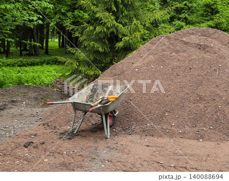 Close up metal wheelbarrow rests beside a pile of crushed red mulch. 140088694