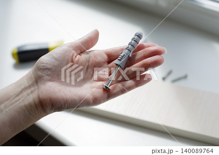 Hand holds a gray plastic dowel near a lighted window. Screwdriver and screws are in the background on a sunny day. Hand holds a gray plastic dowel near a lighted window. Screwdriver and screws are in the background on a sunny day. 140089764