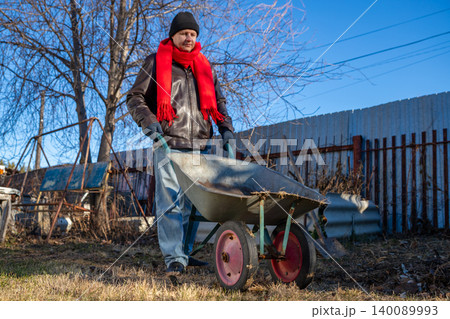 Man gardener pushing wheelbarrow in outdoor garden. Rural lifestyle concept at autumn season. Farmer working on plot near village fence during sunny weather. Active male labor activity. 140089993